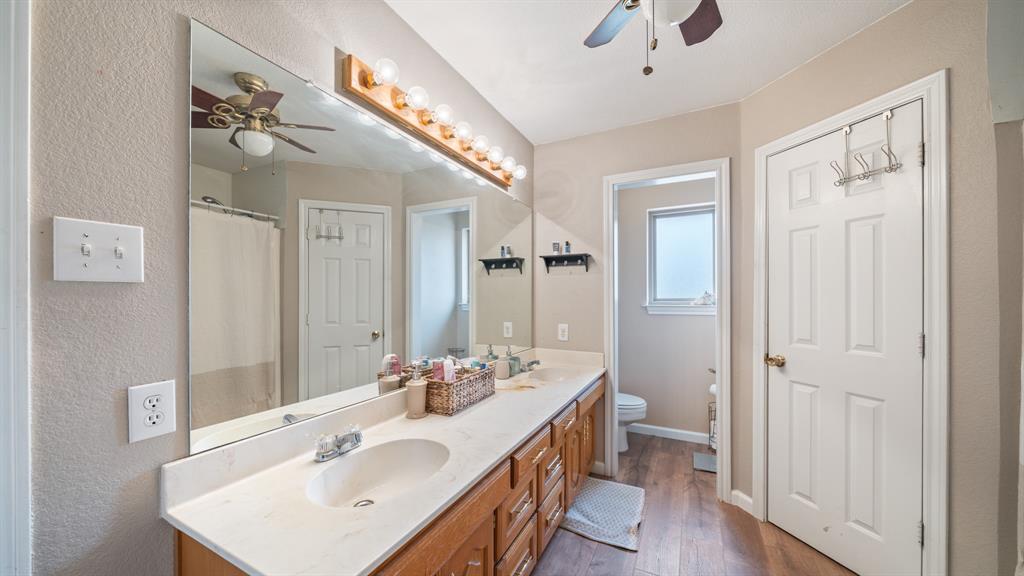 Double vanity featuring a light-toned countertop with integrated dual sinks, wood-finish cabinetry, full-width wall mirror, and wood-finish flooring