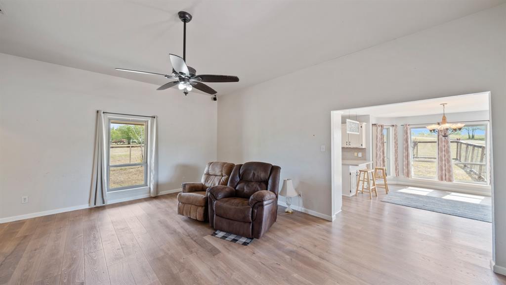 Living area featuring wood-finish flooring, a ceiling fan with integrated lighting, and neutral wall tones, highlighted by natural light through large windows