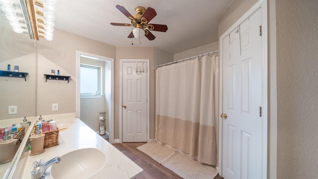 Bathroom featuring a vanity with integrated sink, expansive wall-to-wall mirror, multi-bulb overhead lighting, ceiling fan, and wood-finish flooring