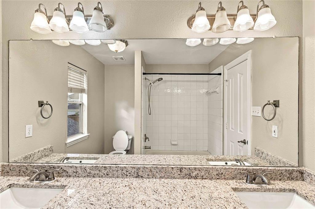 Dual-sink vanity with stone countertop, full-width vanity mirror, dual four-light fixtures, and white tile shower surround with a handheld showerhead