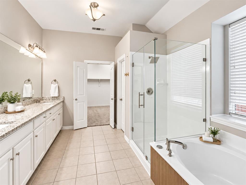 Double vanity with a stone-finish countertop, walk-in glass-enclosed shower, soaking tub with wood-toned surround, ceramic tile flooring, and integrated wall-to-wall mirror