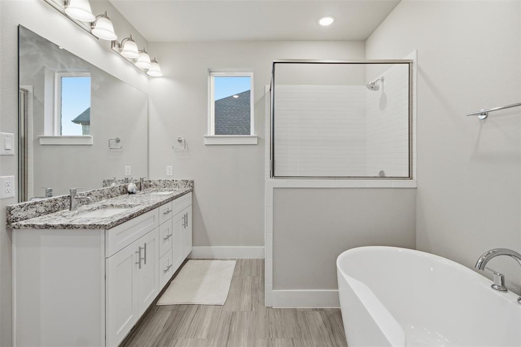Double vanity featuring stone countertops and integrated sinks, accompanied by a freestanding soaking tub, a tiled glass-enclosed shower, and wood-finish flooring