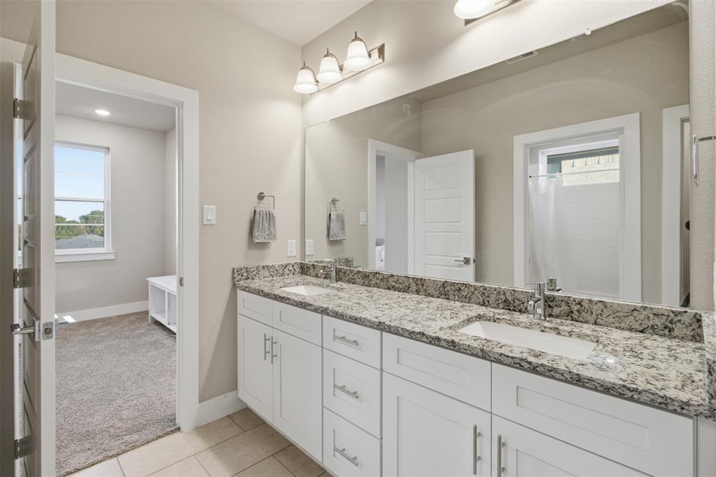 Dual-sink vanity with stone-finish countertop, white shaker-style cabinetry, large wall-mounted mirror, and three-bulb vanity lighting