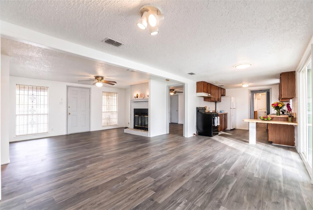 Open-concept interior featuring wood-finish flooring, a central fireplace with a brick surround, a multi-light ceiling fixture, and wood cabinetry Open-concept interior featuring wood-finish flooring, a central fireplace with a brick surround, a multi-light ceiling fixture, and wood cabinetry