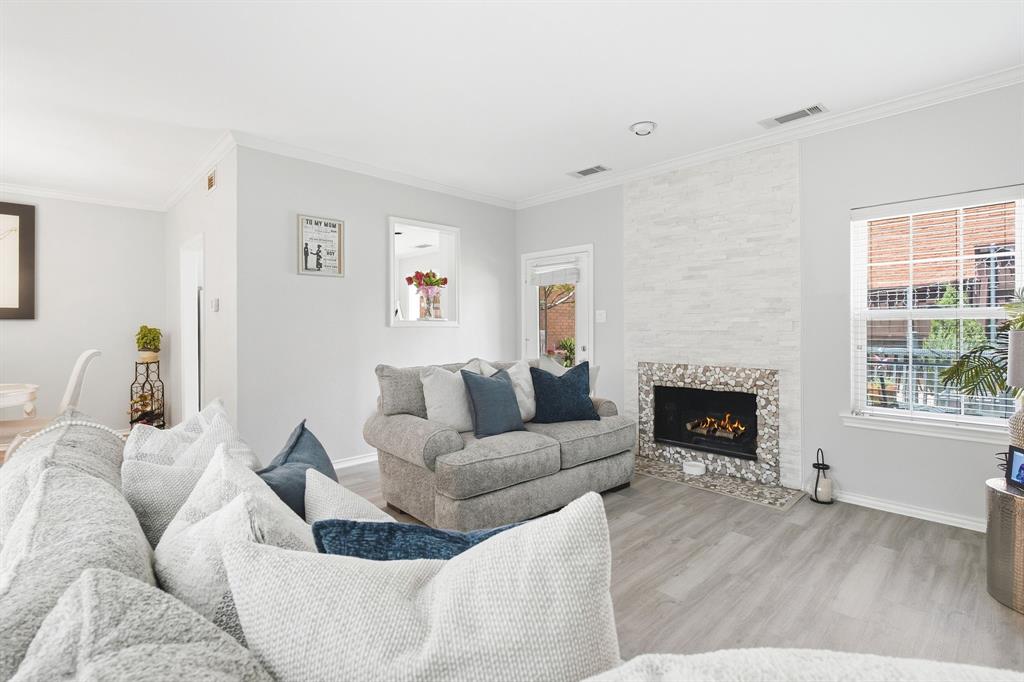 Living area featuring wood-finish flooring, a fireplace with a stacked-stone surround, decorative pebble-tile hearth, and a window with white casing Living area featuring wood-finish flooring, a fireplace with a stacked-stone surround, decorative pebble-tile hearth, and a window with white casing