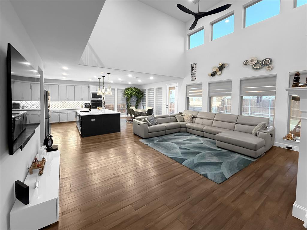 Open-concept interior featuring double-height ceilings, wood-finish flooring, a kitchen island with stone countertops, recessed lighting, and large windows with roller shades