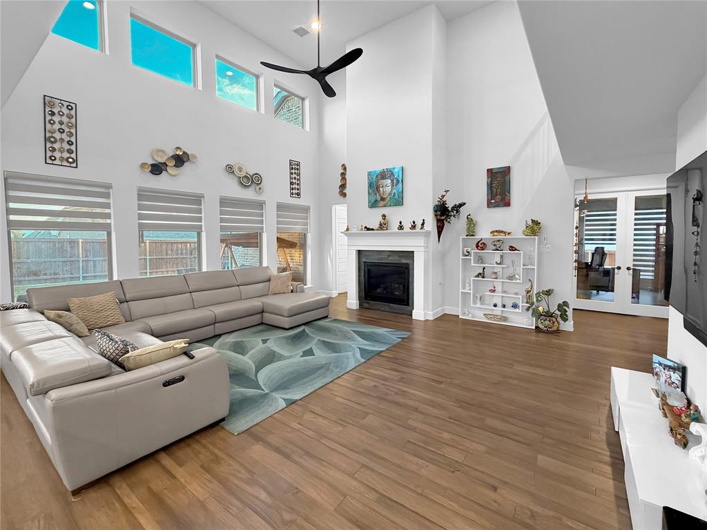 High-ceiling living area featuring wood-finish flooring, a fireplace with a stone-surround mantel, multi-level windows, and a ceiling fan