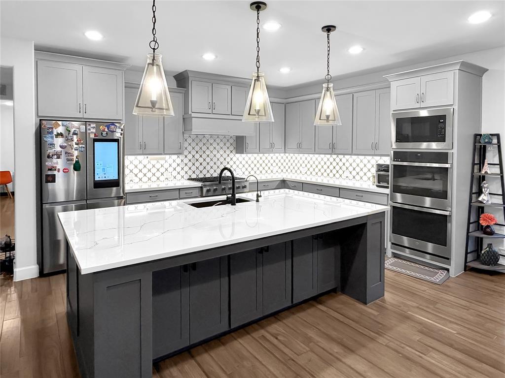 Kitchen featuring a central island with quartz countertop, grey shaker-style cabinetry, stainless steel double wall oven, geometric patterned backsplash, and three pendant lights