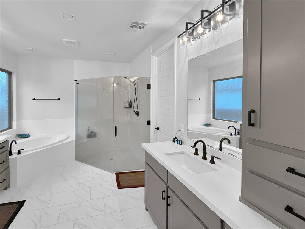 Bathroom featuring a dual-sink vanity with stone-composite countertops, a glass-enclosed walk-in shower, a soaking tub, and tile flooring