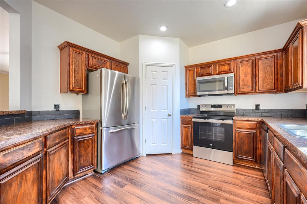 Kitchen featuring wood-finish cabinetry, stainless steel appliances, speckled stone-finish countertops, dark tile backsplash, and wood-finish flooring