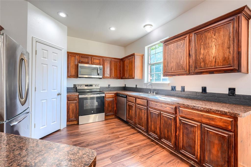 Kitchen featuring rich-toned wood cabinetry, stone-finish countertops, stainless steel appliances, dark tile backsplash, and wood-finish flooring