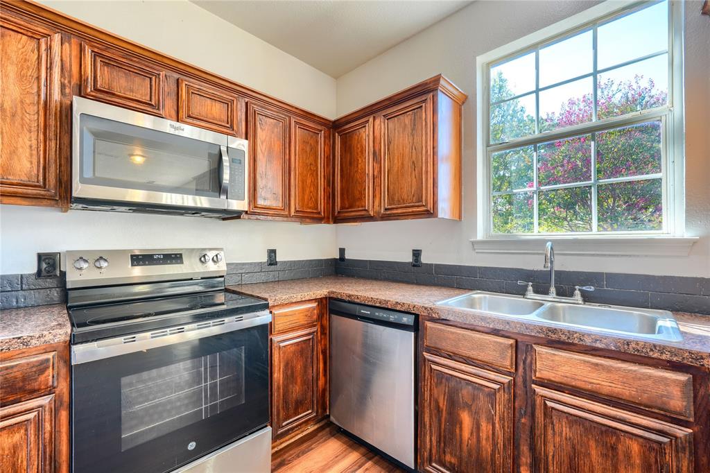 Kitchen featuring stained wood cabinetry, stainless steel appliances, dark tile backsplash, speckled countertops, and a double-basin stainless steel sink under a multi-pane window
