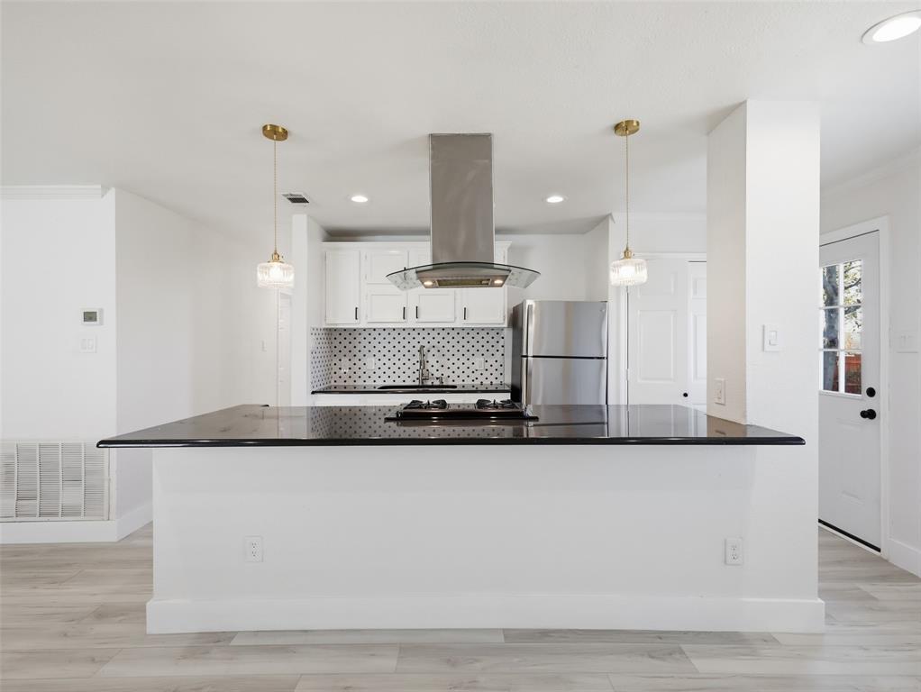 Kitchen featuring a central island with a gas cooktop, dark stone-finish countertop, stainless steel vent hood, white cabinetry, and glass pendant lighting
