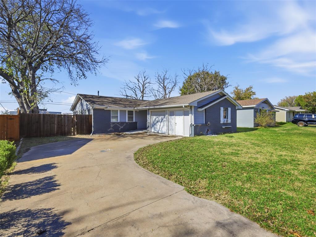 Single-story exterior featuring painted brick siding, an attached two-car garage, and a concrete driveway on a spacious lot with mature trees and wood fencing