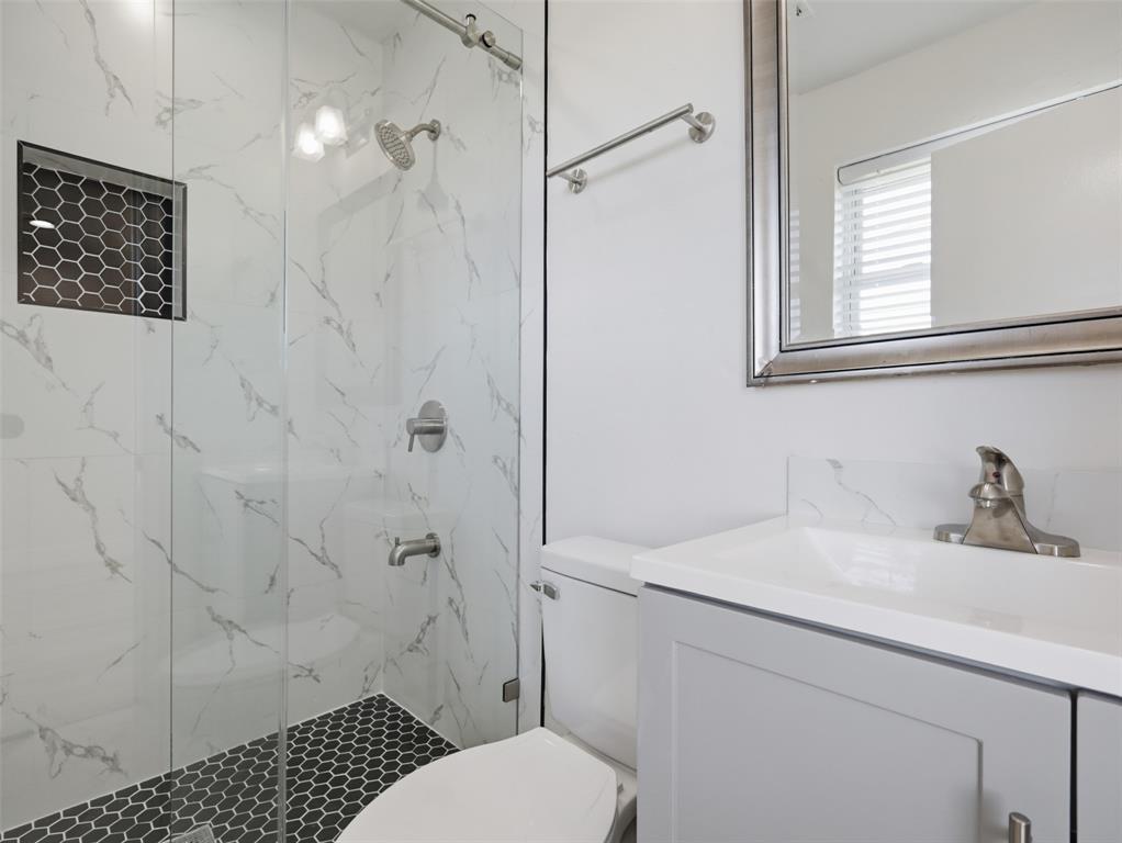 Walk-in shower with stone-finish tile surround, hexagon tile floor, and recessed niche, paired with a gray vanity featuring a rectangular integrated basin and metallic fixtures