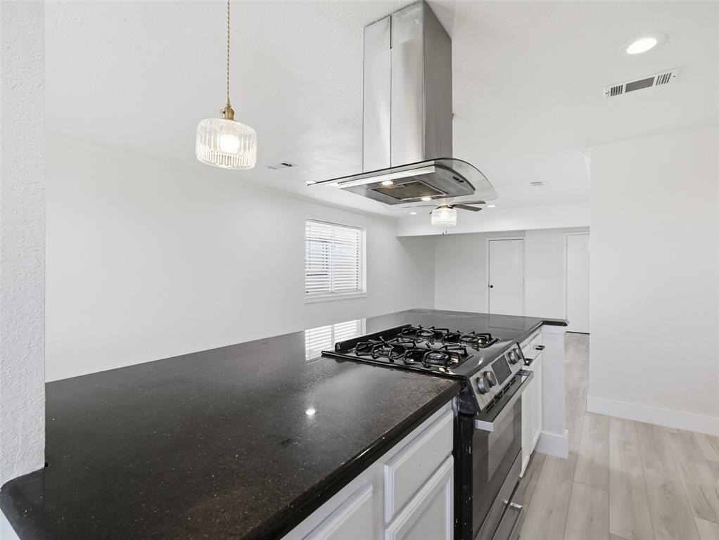 Kitchen featuring a dark stone countertop, a stainless steel gas range with an overhead exhaust hood, wood-finish flooring, and a glass pendant light fixture