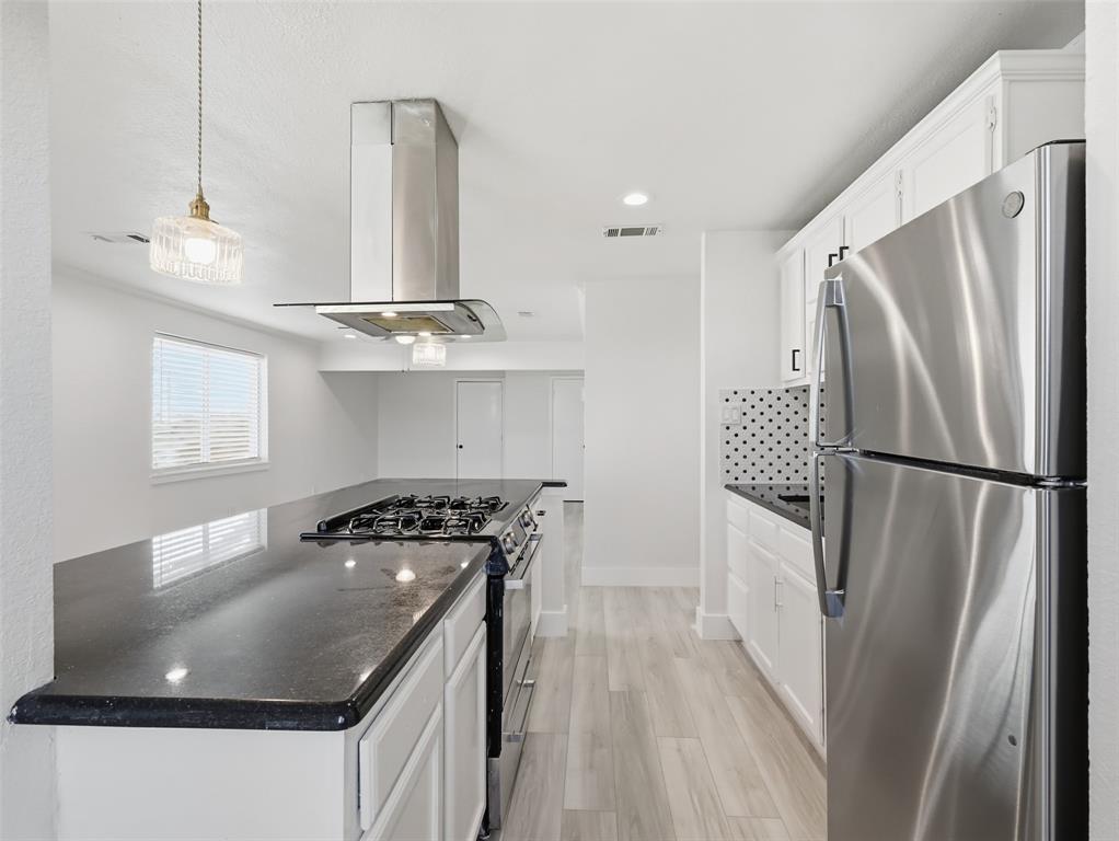 Kitchen featuring white cabinetry, dark stone-finish countertops, stainless steel appliances including a gas range, a modern stainless steel chimney-style range hood, and wood-finish flooring