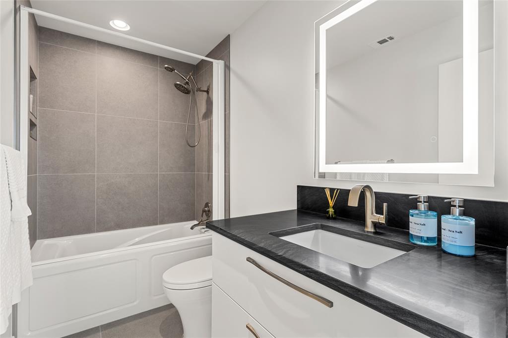 Bathroom featuring a floating white vanity with dark stone-finish countertop, undermount sink, illuminated wall-mounted LED mirror, and a shower-tub combination with retractable privacy door. 24 inch custom tile work.