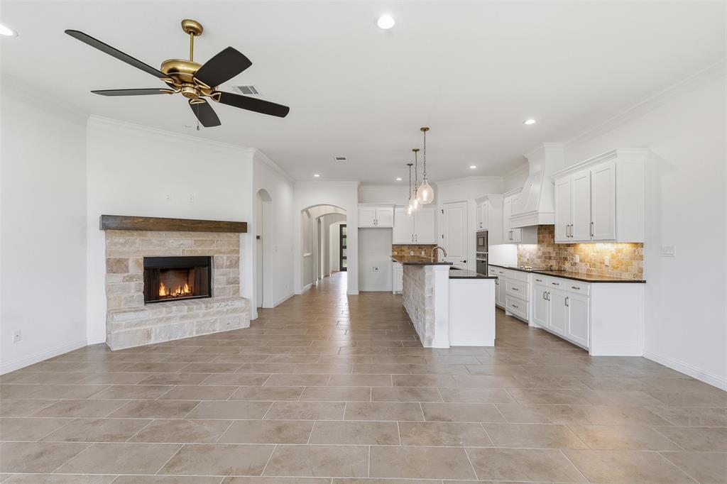 Open-concept interior featuring large-format tile flooring, a floor-to-ceiling stone fireplace with a wood mantel, white cabinetry, dark countertops, and a multi-light pendant fixture over the kitchen island