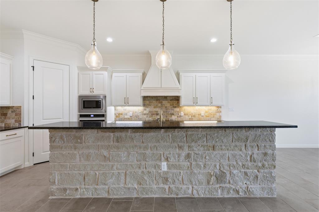 Kitchen featuring a stone-clad island with a dark countertop, white cabinetry, custom range hood, glass pendant lighting, and stone backsplash