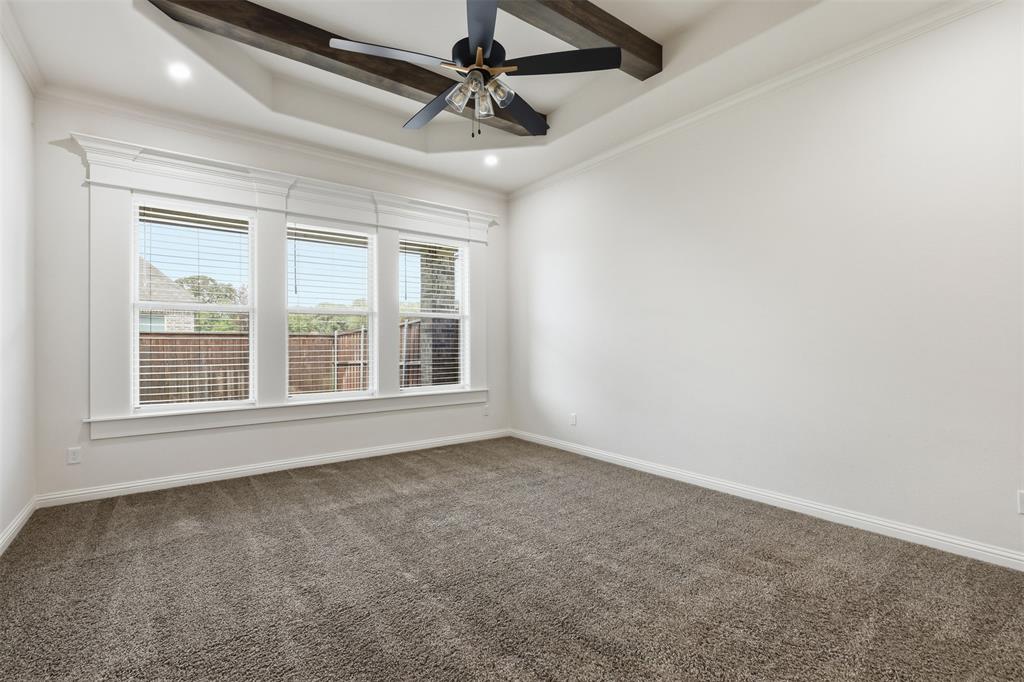 Tray ceiling with exposed wooden beams, center-mounted ceiling fan, neutral wall-to-wall carpeting, and a triple-pane window assembly with detailed millwork trim