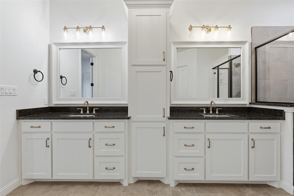 Dual-sink vanity featuring white cabinetry, dark stone-finish countertops, central built-in storage, framed mirrors, and metallic wall-mounted light fixtures