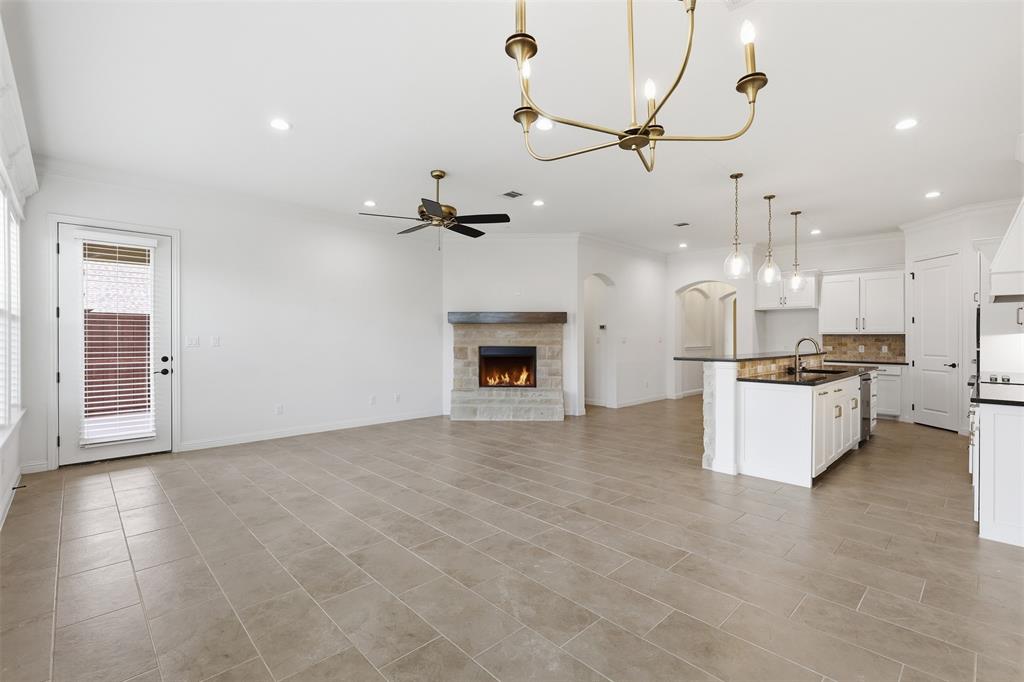 Open concept living area featuring large-format tile flooring, a stone-surround fireplace with a wooden mantel, a kitchen island with an integrated sink, white shaker-style cabinetry, and multi-light chandelier fixtures