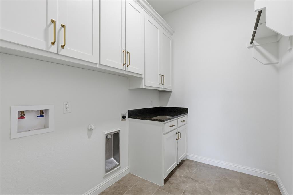 Laundry room featuring white shaker-style cabinetry with brass-tone hardware, a dark stone-surface countertop, an integrated sink, and tile flooring