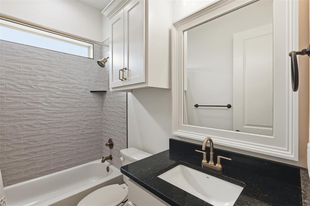 Bathroom featuring a dark stone countertop, integrated sink with a metallic faucet, white cabinetry, a framed mirror, and a tub surround with textured tile