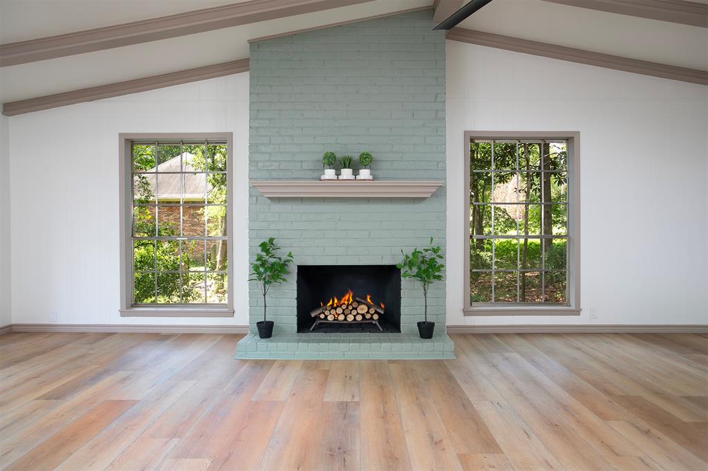 Vaulted ceiling with exposed beams, painted brick fireplace with mantle, wood-finish flooring, and large multi-pane windows
