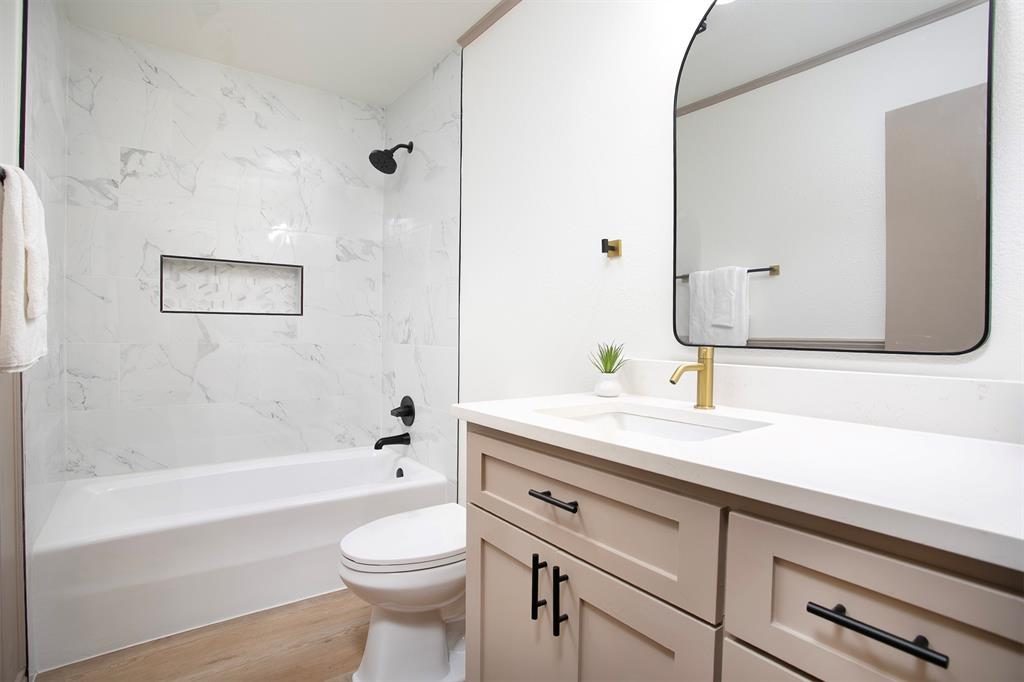 Bathroom featuring a vanity with white countertop, arched wall-mounted mirror, wood-finish flooring, and a tub-shower combination with built-in niche and contrasting fixtures