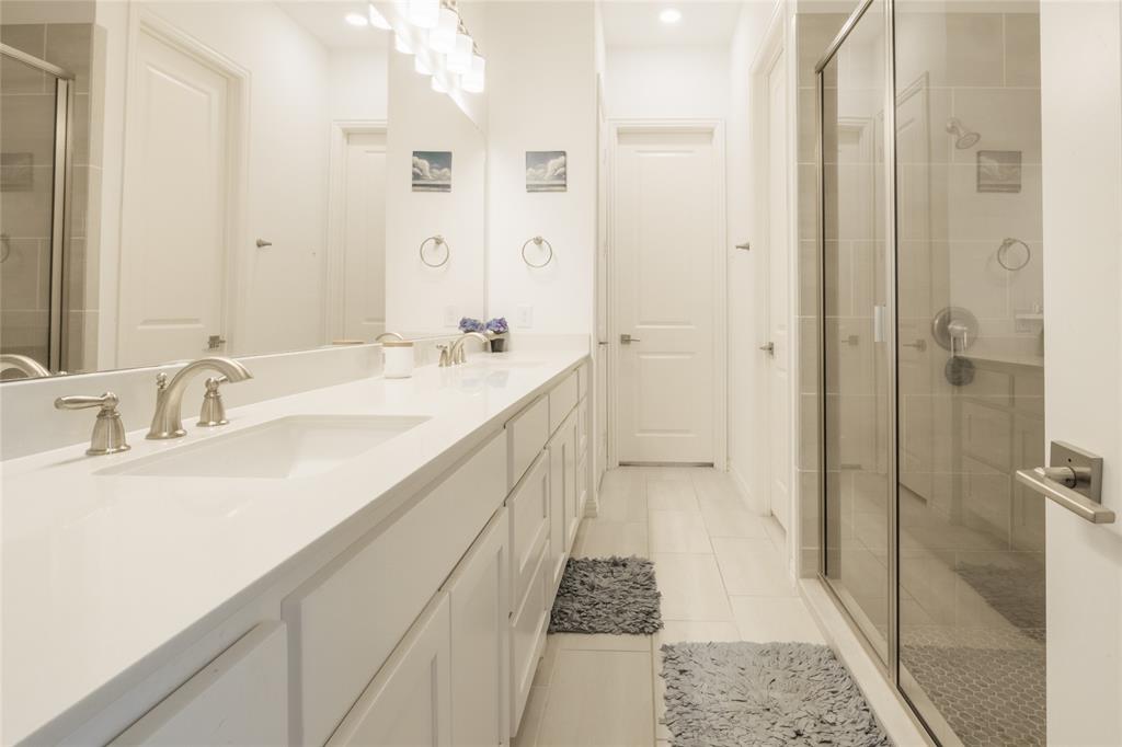 Bathroom featuring a long double vanity with solid surface countertops, dual undermount sinks, white shaker-style cabinetry, and a glass-enclosed shower with tile surround