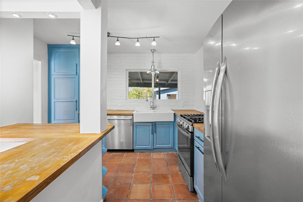 Kitchen featuring a ceramic farmhouse sink, wood-finish countertops, stainless steel appliances, blue cabinetry, and terra-cotta tile flooring
