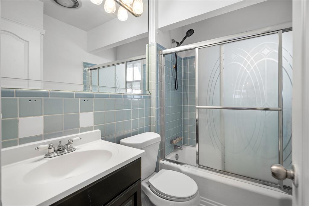 Bathroom featuring a dark-finish vanity with a white integrated sink, full-width wall mirror, shower-tub combination with blue wall tiling, and glass sliding enclosure with etched detailing