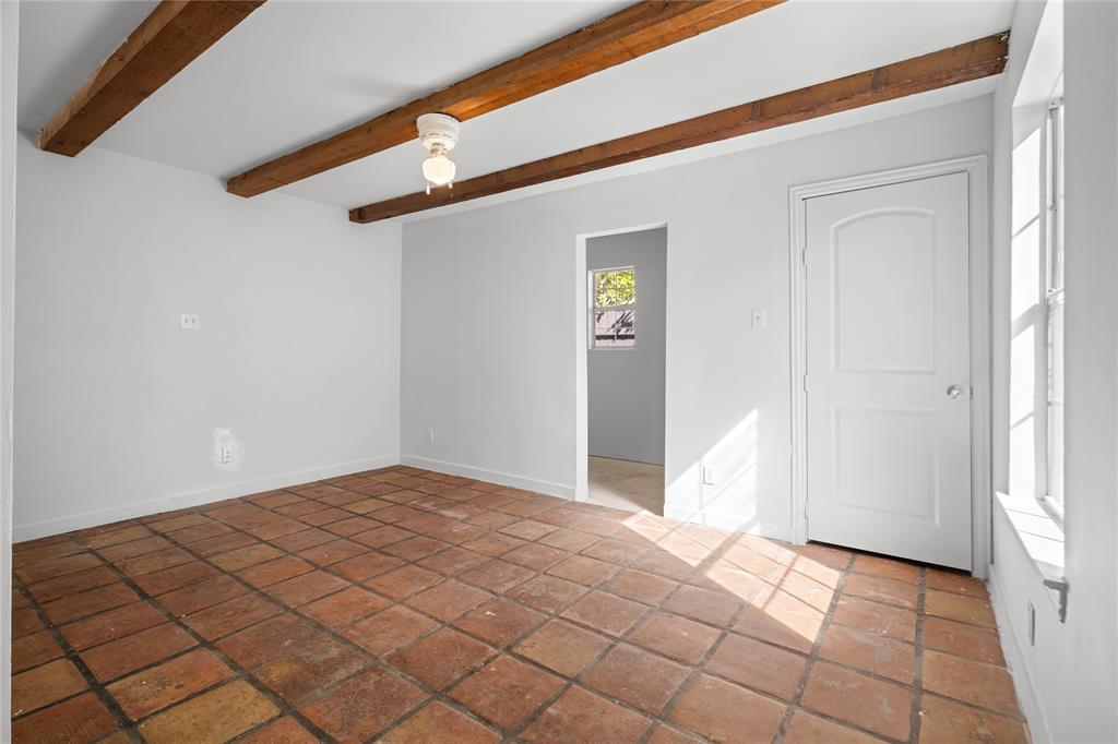 Room featuring exposed wooden ceiling beams, terracotta tile flooring, a multi-pane window, and an arched-panel interior door