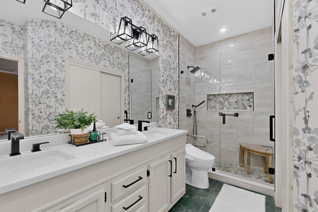 Recently update primary bathroom features dual-sink vanity with a solid-surface countertop and matte black fixtures, walk-in glass-enclosed shower with tiled walls, built-in shower niche, and custom accent wall covering.