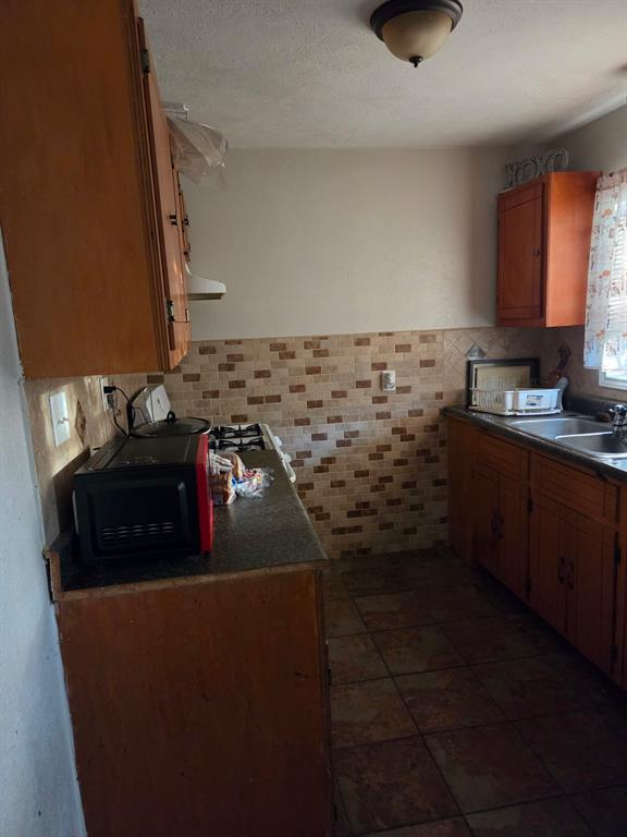 Kitchen featuring stained wood cabinetry, stone tile backsplash, stone-finish floor tile, and a double-basin stainless steel sink Kitchen featuring stained wood cabinetry, stone tile backsplash, stone-finish floor tile, and a double-basin stainless steel sink