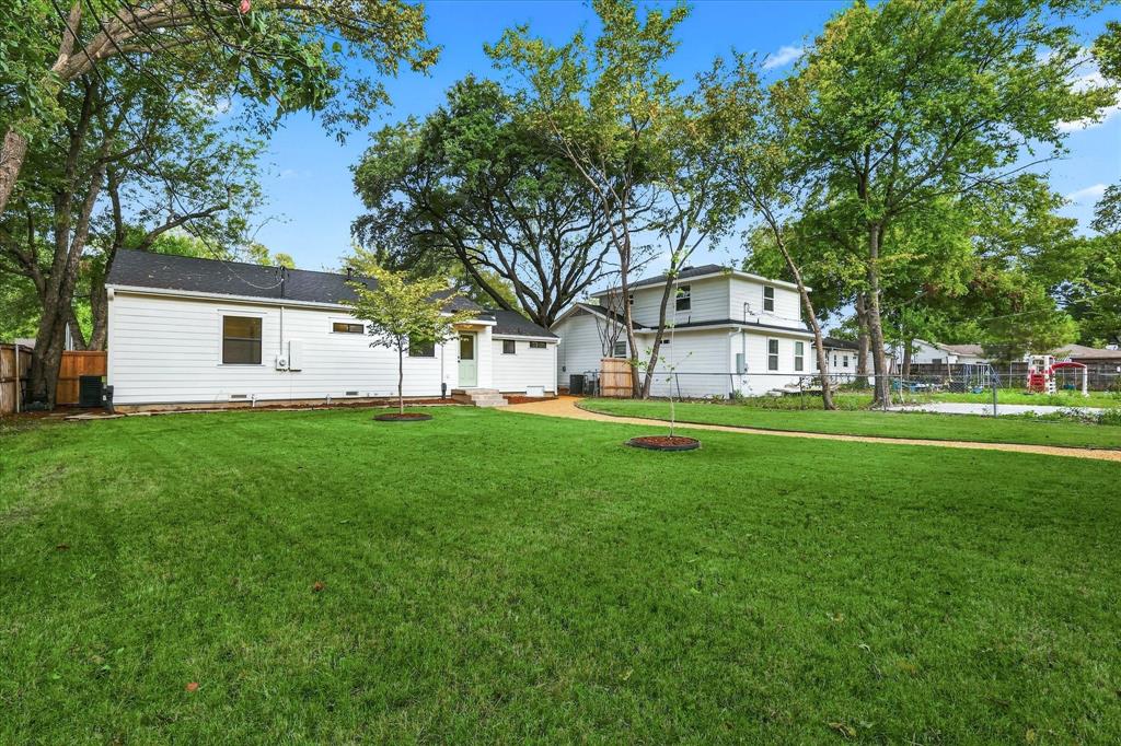 Expansive backyard featuring a large grass lawn, mature shade trees, a light-toned gravel pathway, and white horizontal siding exteriors