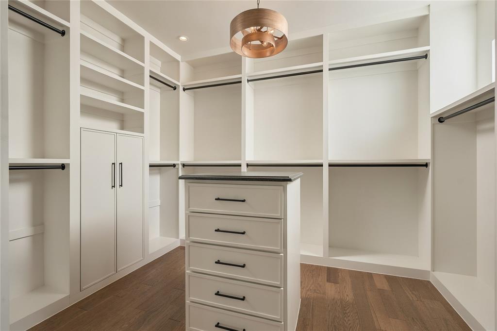 Custom walk-in closet featuring extensive built-in white cabinetry, a center storage island with drawers, dark hardware, wood-finish flooring, and a tiered drum pendant light