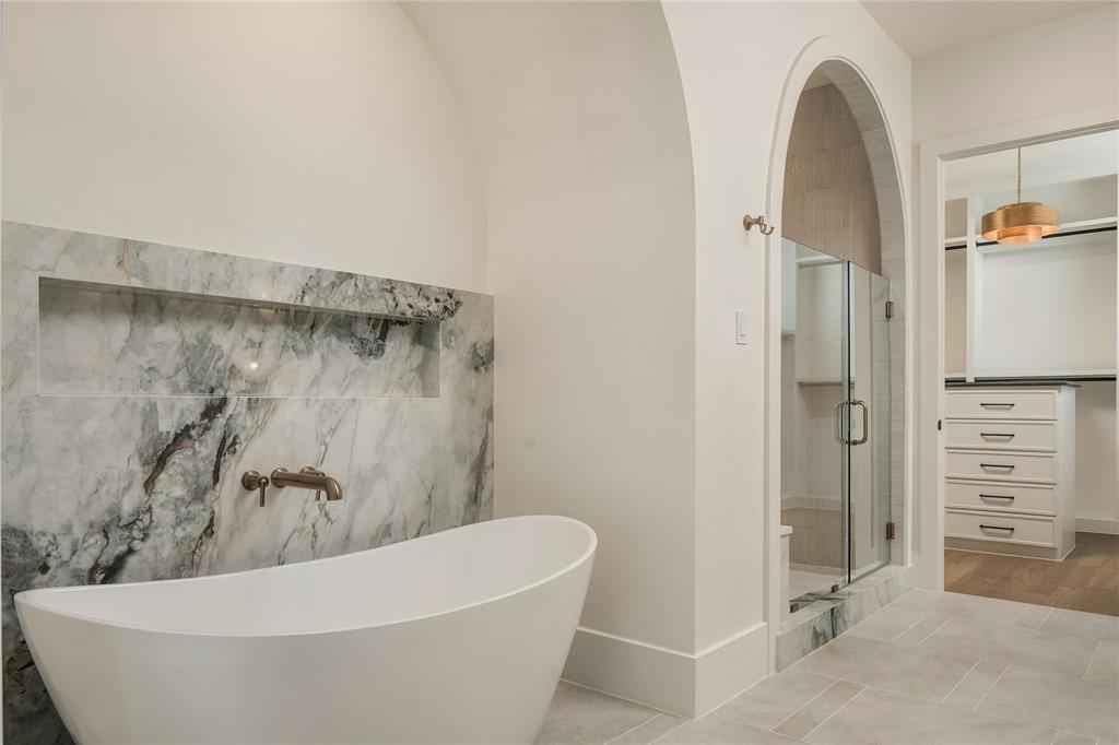 Freestanding soaking tub with wall-mounted faucet, arched shower entry, natural stone wall accent with recessed shelving, and walk-in closet featuring built-in cabinetry