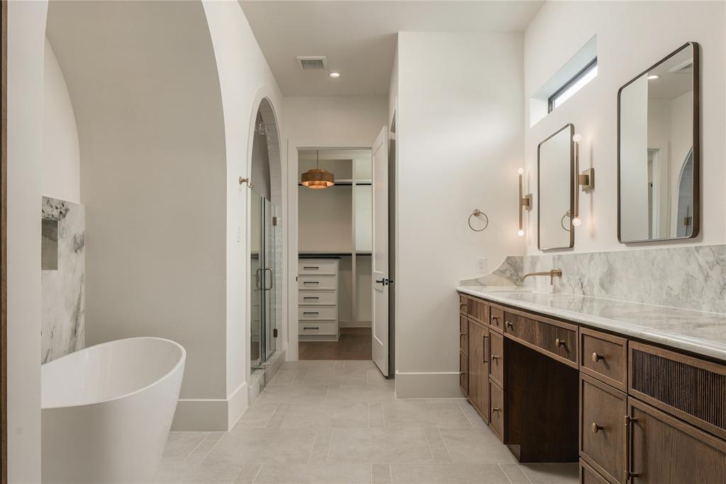 Freestanding soaking tub with a stone-accented alcove, double-vanity cabinetry with stone surfacing, arched walk-in shower, and twin rectangular mirrors with wall-mounted lighting