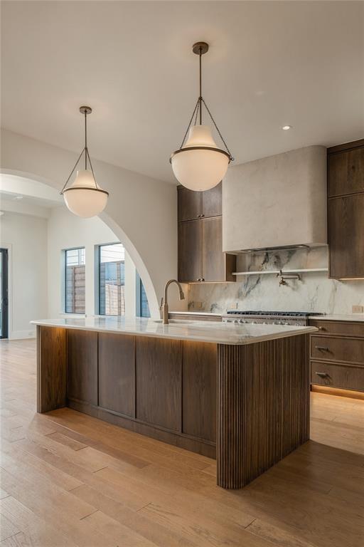 Kitchen featuring a center island with a stone countertop, fluted wood-finish cabinetry, a plaster-finish range hood, stone slab backsplash, and glass pendant lighting