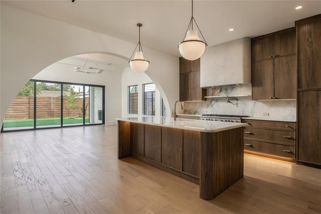 Kitchen featuring custom wood cabinetry, a stone-surround range hood, a large island with integrated under-cabinet lighting, wood-finish flooring, and a prominent architectural archway