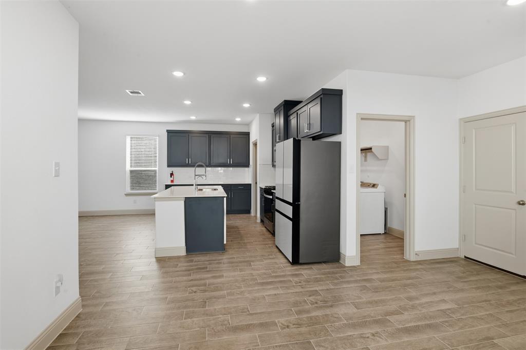 Kitchen featuring dark cabinetry, center island with an integrated sink, neutral stone-finish countertops, wood-finish flooring, and recessed ceiling lighting