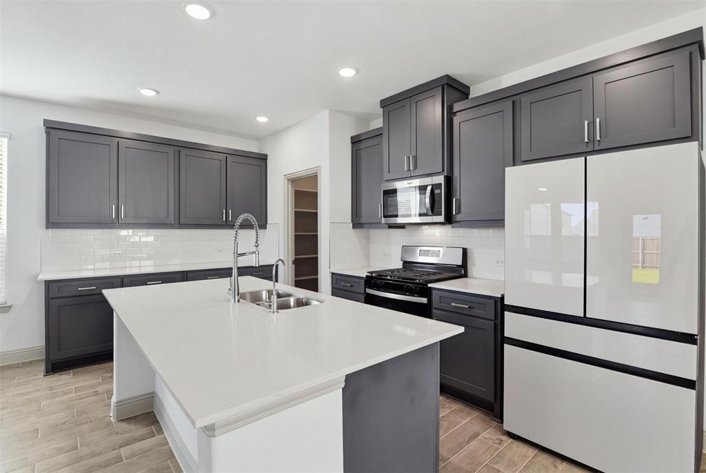 Kitchen featuring dark shaker-style cabinetry, quartz countertops, a central island with an undermount dual-basin sink, white subway tile backsplash, and wood-finish flooring