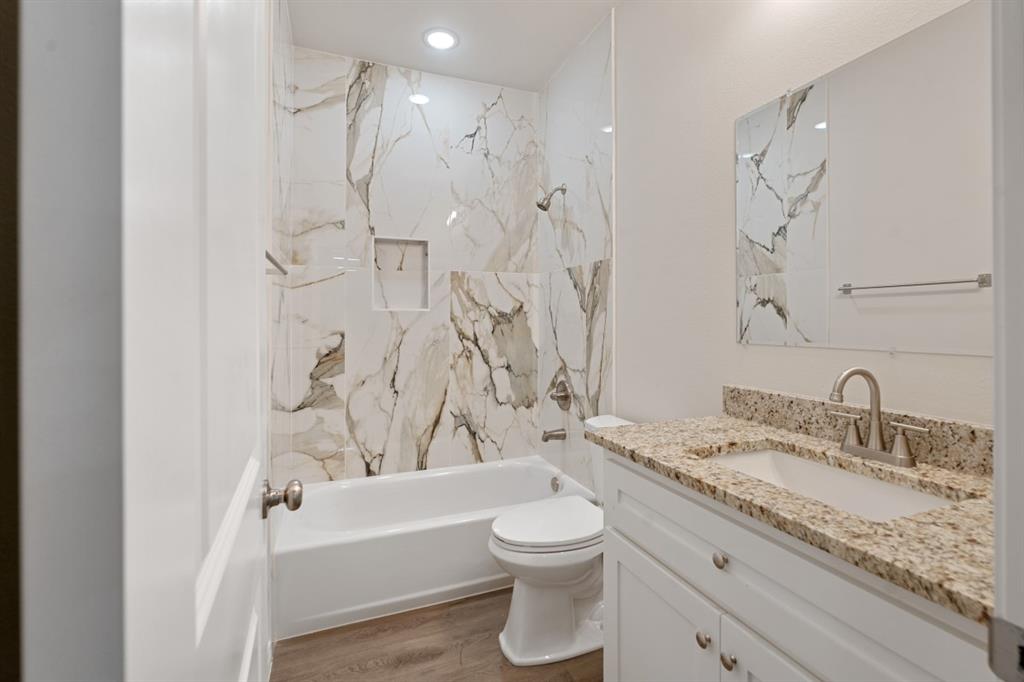 Bathroom featuring a tub-shower combination with floor-to-ceiling stone-pattern tile surround, built-in wall niche, vanity with stone-surface countertop and integrated sink, and wood-finish flooring