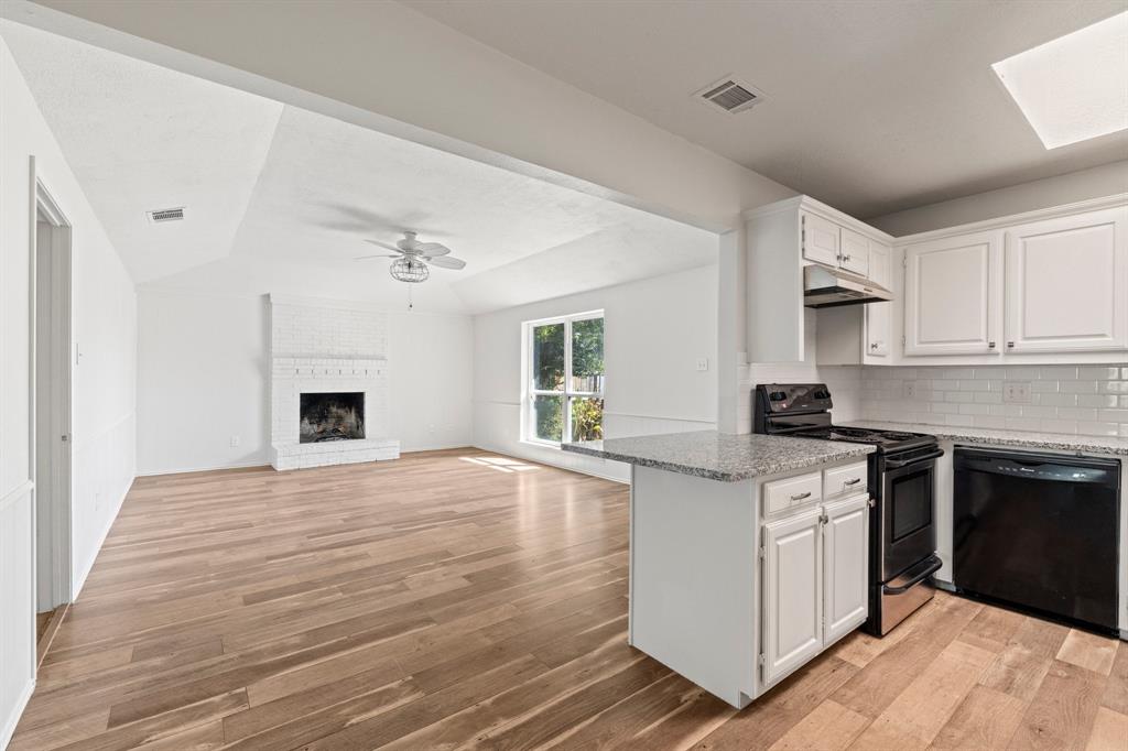 Open-concept interior featuring wood-finish flooring, a white painted brick fireplace, shaker-style cabinetry, stone countertops, and a subway tile backsplash