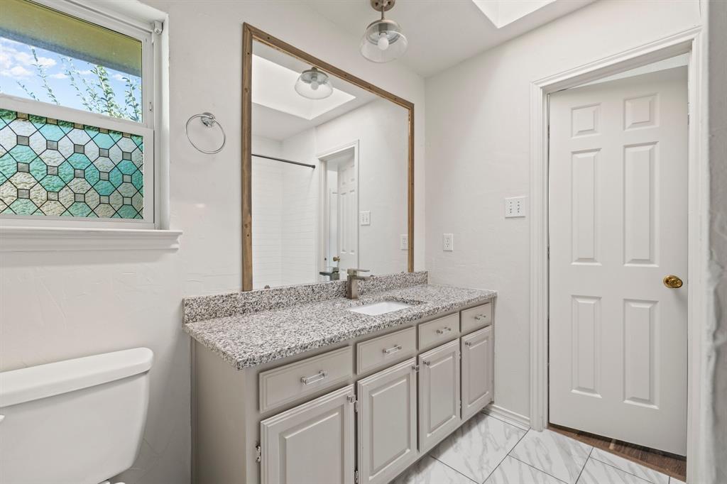 Vanity featuring a stone-look countertop, integrated sink, and shaker-style cabinetry, complemented by a large framed mirror, patterned privacy window glass, and light-toned tile flooring