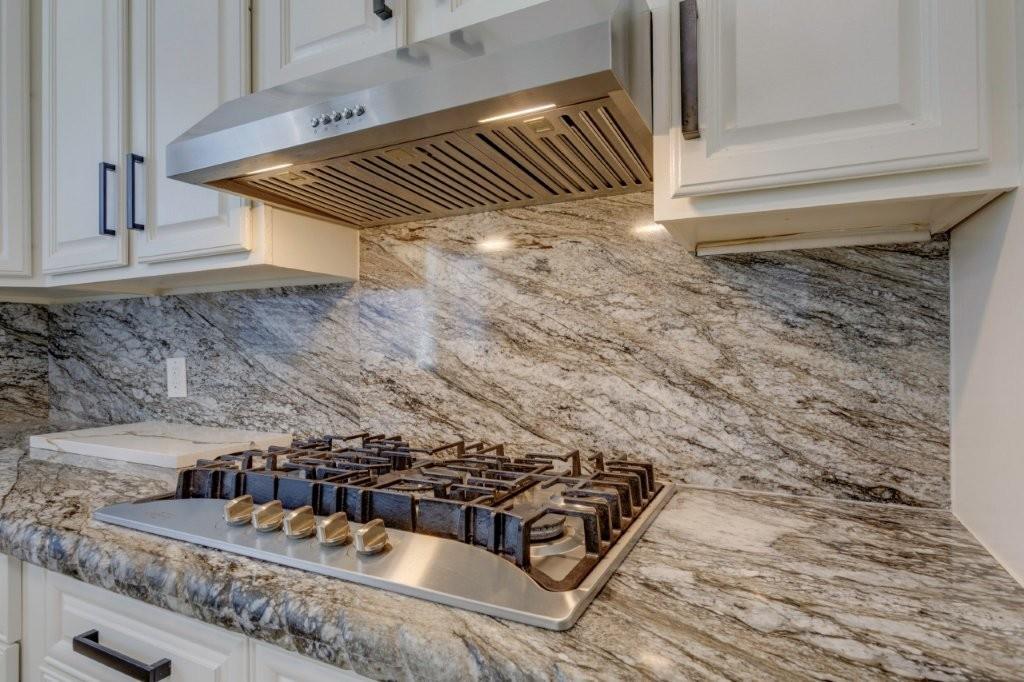 Custom white cabinetry with dark hardware, stone countertop and full-height backsplash, stainless steel gas cooktop, and stainless steel range hood
