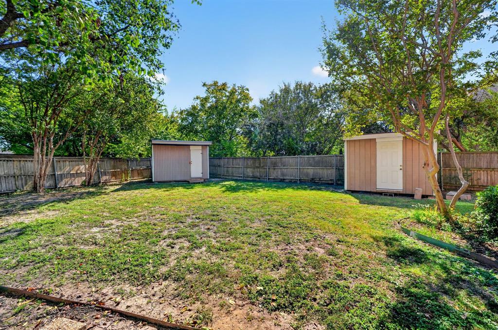 Fenced backyard featuring mature trees, a grass lawn, and two detached storage structures